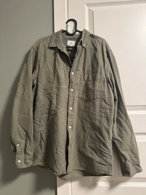 Olivgrön skjorta från H&M - Olivgrön skjorta från H&M i mjuk bomull med klassisk krage och vita knappar. Skjortan har en bröstficka och långa ärmar med knapp vid ärmslut. Perfekt för en avslappnad och stilren look.