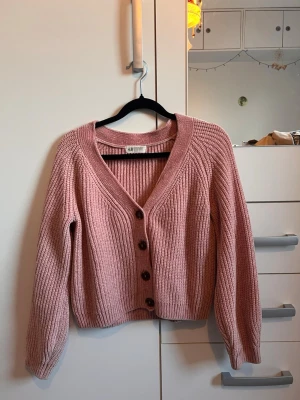 Rosa ribbad kofta från H&M - Mysig rosa kofta från H&M med bred ribbstickning och stora bruna knappar framtill. Koftan har v-ringad hals, långa ärmar och en croppad passform. Perfekt för lager på lager och enkel att styla till olika outfits.