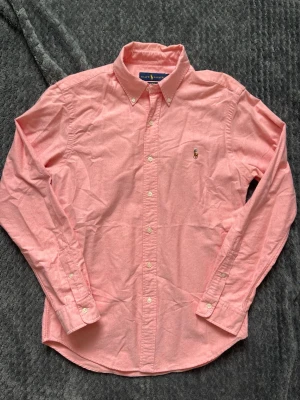 Ralph Lauren skjorta rosa  - Klassisk rosa skjorta från Ralph Lauren med button-down krage och broderad logga på bröstet. Skjortan har långa ärmar, vita knappar och är tillverkad i bomull. Perfekt för en stilren och avslappnad look.
