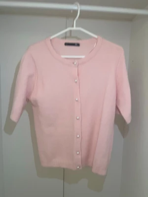 Ljusrosa kofta med pärlknappar - Supersöt ljusrosa kofta med korta ärmar och rund halsringning. Koftan har pärlknappar framtill och är stickad i ett mjukt material. Perfekt för att addera en feminin touch till din outfit.