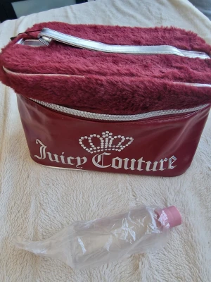Juicy Couture vinröd make up bag  - Juicy Couture handväska i vinrött med fluffig fuskpäls upptill och silvriga dragkedjor. Väskan har ett stort Juicy Couture-tryck med krona framtill och är tillverkad i glansigt konstläder. Perfekt för dig som gillar statement-accessoarer.