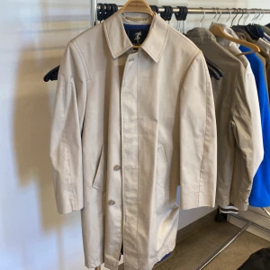 Beige trenchcoat från Rain Wind - Stilren beige trenchcoat med klassisk krage och knäppning framtill. Jackan har två snedställda fickor och ett matchande bälte i midjan. Perfekt för dig som gillar tidlös och enkel stil. 