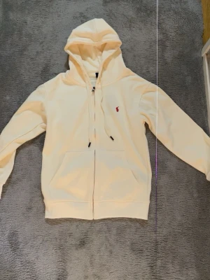 Vit hoodie från Polo Ralph Lauren - Säljer en stilren vit hoodie från Polo Ralph Lauren med dragkedja, huva med dragsko och kängurufickor. Klassisk röd logga broderad på bröstet. Tillverkad i mjukt bomullsmaterial som känns skönt mot huden. Perfekt för en avslappnad och trendig look. Kan sitta Xs