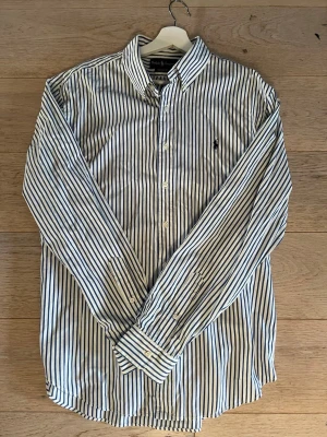 Randig skjorta från Ralph Lauren - Klassisk vit skjorta med blåa ränder från Ralph Lauren. Skjortan har lång ärm, knappar framtill och en broderad logga på bröstet. Tillverkad i bomull och har en tidlös, stilren look med button-down krage.
