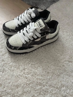 Svarta och vita DAILO & MASM sneakers - Snygga sneakers från DAILO & MASM i svart och vitt med coolt mönster. Skorna har vita snören, perforerad tå och svartvit yttersula. Märkeslogga på plösen och bekväm låg modell som passar till streetwear-stilen.