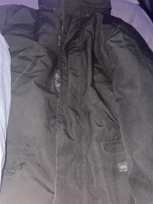 Svart parkas från Canada Goose - Säljer en svart parkas från Canada Goose med klassisk patch på ärmen. Jackan har huva, dragkedja och flera fickor framtill dragkedjan är dock trasig . Perfekt för kalla dagar med sin rejäla och stilrena design. Materialet är slitstarkt och håller värmen bra.storlek m