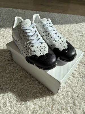 MaisonMargiela gats sneakers med svart splash - Unika vita sneakers från Nike med svart splash-design över tån och svarta färgstänk över hela ovandelen. Klassisk låg modell med vita skosnören och diskret Nike-logga på hälen. Tillverkade i läder för en clean och modern look.
