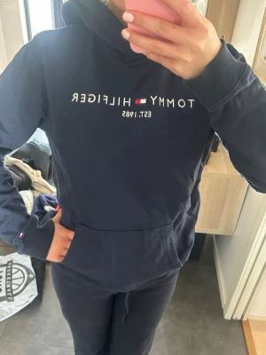 Marinblå hoodie från Tommy Hilfiger - Snygg marinblå hoodie från Tommy Hilfiger med klassisk logga och texten 'EST. 1985' på bröstet. Hoodien har huva, magficka och ribbade muddar vid ärmar och nederkant. Perfekt för en avslappnad och stilren look.
