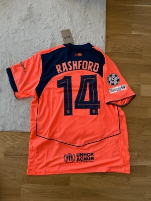 Barcelona Rashford matchtröja orange - Snygg Barcelona matchtröja i orange med mörkblå detaljer och tryck. Rashford och nummer 14 på ryggen, Champions League-märke på ärmen samt UNHCR och Spotify-loggor. Tillverkad i lätt och ventilerande material, perfekt för fotboll eller träning.