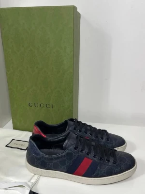 Gucci ace sneakers med blå monogram - Snygga sneakers från Gucci i mörkblått tyg med klassiskt GG-monogram. Skorna har svarta snören, vit sula och de ikoniska röda och blå ränderna på sidan. Baksidan har en röd detalj i läder. Perfekta för dig som vill ha en exklusiv och trendig look.