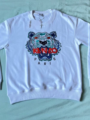 Vit Kenzo sweatshirt med tiger - Vit sweatshirt från Kenzo med ikonisk broderad tiger på bröstet och texten 'KENZO PARIS' i rött och svart. Tröjan har rund halsringning och ribbade muddar. Materialet är mjukt och bekvämt, perfekt för en cool och avslappnad look.