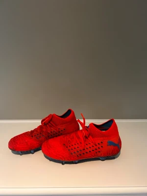 Röda Puma fotbollsskor med blå detaljer - Säljer ett par röda fotbollsskor från Puma med blå logga och blåmönstrad sula. Skorna har snörning, stickad ovandel och små svarta detaljer på sidorna. Perfekta för dig som vill sticka ut på planen och ha bra grepp på konstgräs eller gräs.