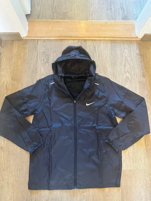 Nike softshell jacka ny utan tags  - Snygg svart vindjacka från Nike med dragkedja framtill och huva. Jackan har två sidofickor, vita detaljer och Nike-logga på bröstet. Tillverkad i lätt och tåligt material som står emot vind. Perfekt för dig som vill ha en sportig och stilren look. Ny skick 