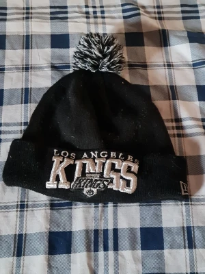 Svart LA Kings mössa med tofs - Säljer en svart stickad mössa från New Era med Los Angeles Kings broderat framtill och en svartvit tofs på toppen. Perfekt för dig som gillar hockey och vill ha en snygg accessoar med sportig vibe.