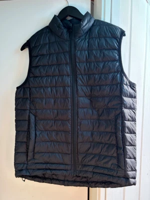 Svart dunväst från Everest - Snygg svart dunväst från Everest med quiltad design och hög krage. Västen har dragkedja framtill och två sidofickor. Perfekt att slänga över en hoodie för extra värme. Lätt och smidig att bära, passar till många olika outfits.