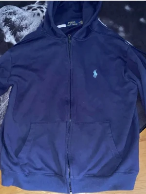 Marinblå hoodie från Polo Ralph Lauren - Snygg marinblå hoodie från Polo Ralph Lauren med dragkedja och klassisk logga broderad på bröstet. Modellen har huva, två fickor framtill och ribbade muddar. Perfekt för en chill och stilren look. Materialet är mjukt och skönt bomullstyg. Det är storlek M men sitter som en S 