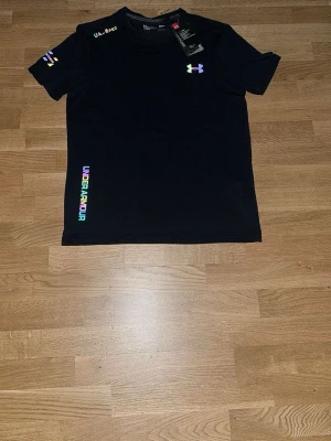 Svart Under Armour t-shirt med färgglada detaljer - Svart t-shirt från Under Armour med färgglada loggor och textdetaljer på bröstet, ärmen och längs sidan. Klassisk passform och rund halsringning. Perfekt för en sportig och avslappnad stil.    