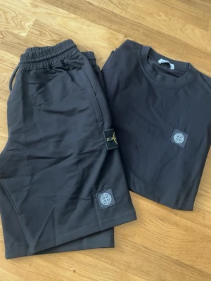 Svart set från Stone Island - Snyggt svart set från Stone Island med matchande t-shirt och shorts. Båda plaggen har den ikoniska Stone Island-loggan framtill. Setet är gjort i mjukt bomullsmaterial och har en avslappnad passform. Perfekt för en stilren och bekväm look.