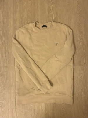 Beige sweatshirt från GANT - Snygg beige sweatshirt från GANT med rund halsringning och broderad logotyp på bröstet. Tröjan har långa ärmar och ribbade muddar vid ärmslut och nederkant. Perfekt för en avslappnad och stilren look.