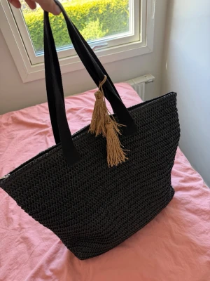 Svart Strandväska med tofsar - Snygg svart strandväska i flätat material med två svarta handtag och dekorativa beige tofsar. Perfekt storlek för att få plats med allt du behöver. Enkel och stilren design som passar till många outfits. Aldrig använd!