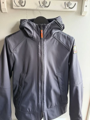 Mörkgrå vindjacka från Parajumpers - Snygg mörkgrå vindjacka från Parajumpers med huva och dragkedja framtill. Jackan har två fickor med dragkedja och en ikonisk PJS-patch på ärmen. Perfekt för blåsiga dagar och har en sportig, stilren look.