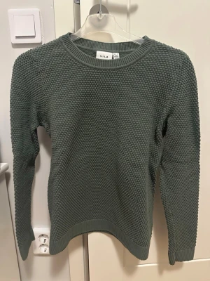 Grön stickad tröja från Vila, motsvarar stl S - Mörkgrön stickad tröja från Vila med rund halsringning och grovstickad struktur. Tröjan har långa ärmar och ribbade muddar vid ärmslut och nederkant. Perfekt för lager-på-lager och enkel att matcha med jeans eller kjol. Liten i storleken så skriver stl S. Använd 1 gång. 