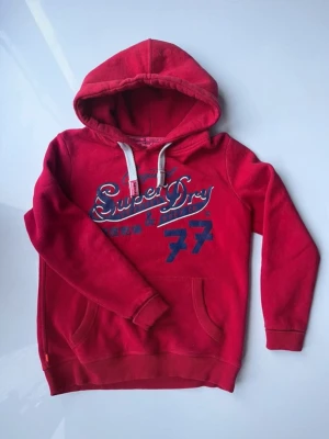 Röd hoodie från Superdry - Snygg röd hoodie från Superdry med blått tryck och vita dragsnören. Klassisk känguruficka framtill och stor huva. Mjuk bomullsblandning som känns skön mot huden. Perfekt för en avslappnad streetwear-look.