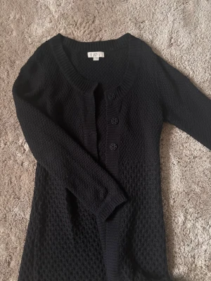 Svart stickad kofta med knappar - Säljer en svart stickad kofta med tre knappar framtill och rund halsringning. Koftan har ett diskret mönster i stickningen och långa ärmar. Perfekt att slänga över en topp för en chill look.