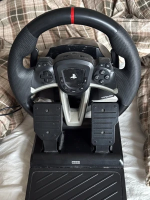 Svart ratt och pedaler från Hori - Säljer en svart racingratt med pedaler från Hori, designad för PlayStation. Ratten har gummerat grepp, PlayStation-knappar och en röd markering upptill. Pedalerna är i svart plast och monterade på en stadig bas. Perfekt för racing- och bilspel. Fungerar till Ps4/ Ps5/ PC
