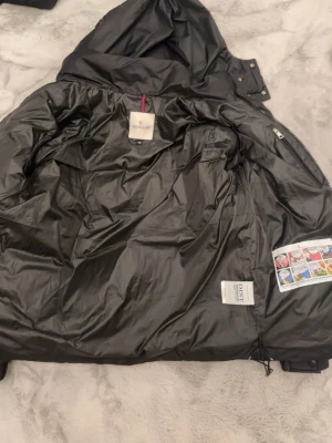 Svart dunjacka från Moncler - Säljer en svart dunjacka från Moncler med avtagbar huva och klassiskt quiltat mönster. Jackan har dragkedja, tryckknappar och Moncler-logga på ärmen. Materialet är glansigt och fyllningen är dun, perfekt för kalla vinterdagar. Använd 1 gång 