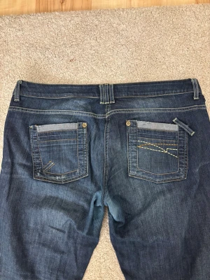 Mörkblå bootcut jeans med detaljer - Säljer ett par mörkblå bootcut jeans med snygga kontrastsömmar och unika detaljer på bakfickorna. Materialet är denim i bomull med en skön känsla. Perfekta för dig som gillar en avslappnad men ändå stilren look. Slitna i botten av byxbenet och lite nötta i grenen. 