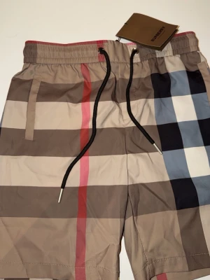 Rutiga badbyxor från Burberry - Säljer ett par snygga badbyxor från Burberry med klassiskt rutmönster i beige, svart, vitt, rött och blått. De har svarta dragsnören, sidofickor och en bakficka. Perfekta för stranden eller poolen och ger en lyxig känsla.