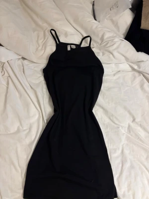 Svart kort klänning med smala band - En enkel och stilren svart kort klänning med smala axelband. Klänningen har en figurnära passform och är perfekt för dig som gillar minimalistisk stil. Tillverkad i mjukt material som känns skönt mot huden.