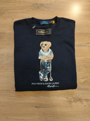 Mörkblå Polo Bear sweatshirt - Mörkblå sweatshirt från Ralph Lauren med ikoniska Polo Bear-trycket framtill. Tröjan har rund halsringning, långa ärmar och ribbade muddar vid ärmslut och nederkant. Perfekt för en avslappnad och trendig stil. Det står att den är Large men den är i storleken Small