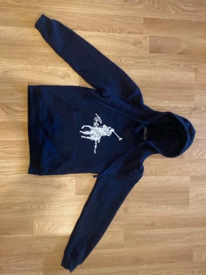 Marinblå hoodie från Polo Ralph Lauren - Snygg marinblå hoodie från Polo Ralph Lauren med stor vit polospelare tryckt på bröstet. Klassisk känguruficka framtill och justerbar huva med dragsko. Tillverkad i mjuk bomullsblandning som känns skön mot huden. Perfekt för en avslappnad och trendig look.