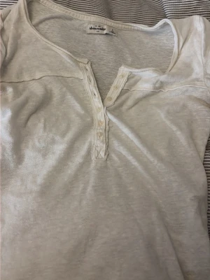 Vit topp med knappar från Abercrombie - Vit topp från Abercrombie med liten v-ringning och dekorativa knappar framtill. Tillverkad i mjukt material och har en enkel, klassisk design som passar till det mesta. Perfekt basplagg för din garderob.