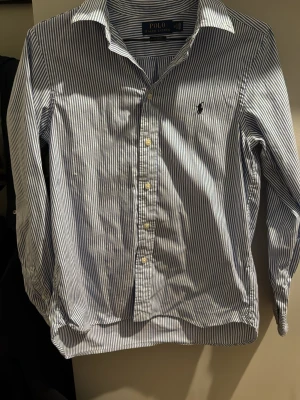 Randig skjorta från Polo Ralph Lauren - Klassisk blåvit randig skjorta från Polo Ralph Lauren i slim fit-modell. Skjortan har långa ärmar, knappar framtill och den ikoniska broderade loggan på bröstet. Tillverkad i bomull och passar perfekt för en stilren look. Använd men fortfarande i bra skick utan defekter. Storleken är barnstorlek men passar xs. Storlek kan även ses på sista bilden!