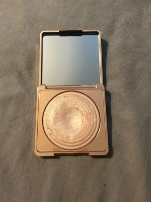 Champagnefärgad highlighter i dosa - Säljer en highlighter i en fyrkantig, guldig dosa med spegel. Produkten har en skimrande champagnefärg och pressad design i cirkulära mönster. Perfekt för att ge glow till ansiktet. Dosan är smidig att ta med i väskan.