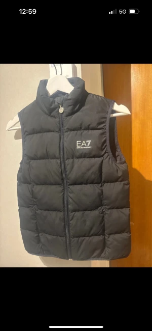 Svart dunväst från EA7 Emporio Armani - Snygg svart dunväst från EA7 Emporio Armani med hög krage och dragkedja framtill. Västen har ett stilrent tryck med logga på bröstet och quiltad design som ger en modern look. Perfekt att slänga över en hoodie för extra värme.