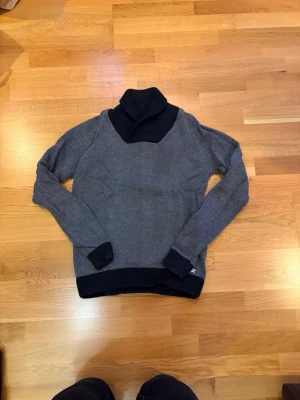 Grå stickad polotröja med svarta detaljer - Snygg grå stickad polotröja med bred svart polokrage och svarta muddar. Tröjan har lång ärm och ribbstickade avslut vid ärmslut och nederkant. Perfekt för kalla dagar och enkel att matcha med jeans eller kjol.