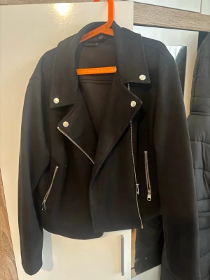 Svart biker skinnjacka med dragkedjor - Snygg svart skinnjacka i bikerstil med silvriga dragkedjor och tryckknappar. Jackan har bred krage och två snedställda fickor med dragkedja. Perfekt för en cool och stilren look.