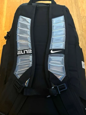 Nike Elite svart ryggsäck - Priset är förhandlingsbart vid snabb affär! ☺️🙌🌟Säljer en svart Nike Elite ryggsäck med unika axelband som har transparenta detaljer. Ryggsäcken har flera fack, meshficka på insidan och justerbara remmar. Perfekt för sport och skola, snygg och funktionell design.