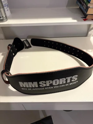 Svart träningsbälte från MM Sports - Snyggt och robust svart träningsbälte från MM Sports med vit logga och texten 'WHY BE AVERAGE WHEN YOU CAN BE MORE'. Bältet är gjort i skinn och har flera hål för justerbar passform. Perfekt för dig som vill ha extra stöd vid gymmet.
