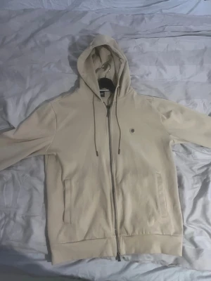 Beige zip hoodie från BOSS - Snygg beige hoodie från BOSS med dragkedja framtill och justerbar huva med snören. Hoodien har två fickor på sidorna och en liten BOSS-logga på bröstet. Perfekt för en chill och stilren look.