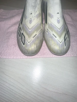Adidas F50 vita fotbollsskor stl 40 - Säljer ett par Adidas F50 fotbollsskor i vitt med svarta och lila detaljer samt guldiga sulor. Skorna har en strömlinjeformad design och är tillverkade i syntetmaterial för lätthet och snabbhet på planen. Perfekta för dig som vill sticka ut med stil och prestanda.