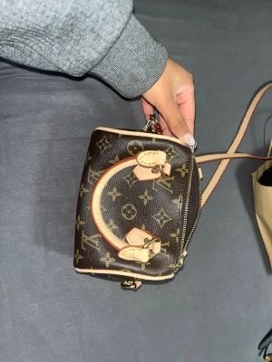 Louis Vuitton Monogram Mini Handväska fake! - Säljer en liten brun handväska från Louis Vuitton med klassiskt monogrammönster och beigea detaljer. Väskan har två handtag, avtagbar axelrem och guldfärgade metalldetaljer. Perfekt för dig som gillar lyxiga accessoarer och vill ha något unikt.