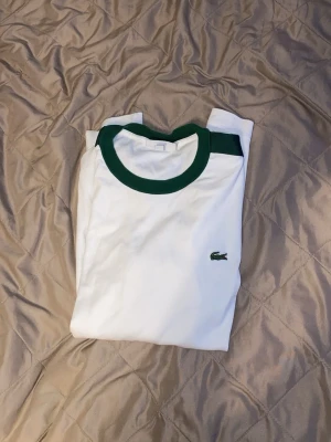 Vit t-shirt med gröna detaljer Lacoste - Säljer en clean vit t-shirt från Lacoste med gröna detaljer runt kragen och axlarna. Klassisk krokodil-logga på bröstet. Perfekt till jeans eller shorts för en sportig vibe. Tillverkad i mjuk bomull för skön känsla hela dagen.