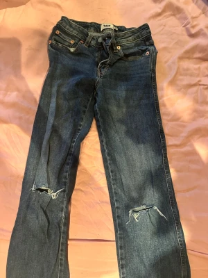 Blå slitna jeans från 157 - Säljer ett par blå jeans från 157 med raka ben och slitningar vid knäna för en trendig look. Jeansen har klassiska fem fickor, knappgylf och detaljerade bakfickor med knappar. Perfekta för dig som gillar en avslappnad och cool stil.