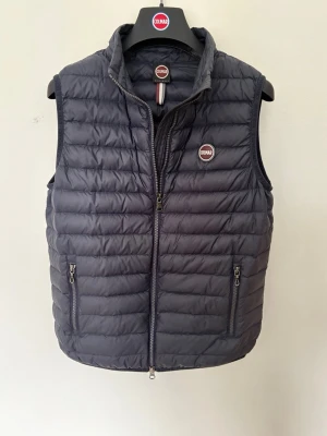 Mörkblå dunväst från Colmar - Snygg mörkblå dunväst från Colmar med quiltad design och hög krage. Västen har två dragkedjeförsedda fickor framtill och en diskret logga på bröstet. Perfekt att slänga över en hoodie eller tröja för extra värme. Materialet är lätt och smidigt. Du får även med en galge från Colmar som är riktigt snygg att ha i rummet!