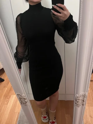 Svart klänning med puffärmar - Snygg svart kortklänning med höghalsad design och transparenta puffärmar i mesh. Klänningen har en tight passform och är perfekt för dig som vill sticka ut med stilrena detaljer. Materialet är stretchigt och bekvämt.